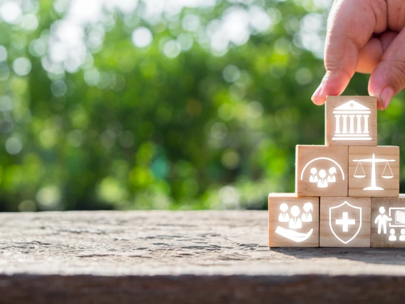 A hand places a wooden block with a government building icon on top of a pyramid of blocks featuring symbols for community, justice, healthcare, education, and support services, representing societal foundations.